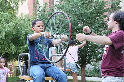 At the festival, King teaches children the conservation of angular momentum by sitting them on a chair on a free-moving pedestal and having them hold a bike wheel vertically. He then gets the tire spinning fast and tells the unaware youngsters to tilt it.
